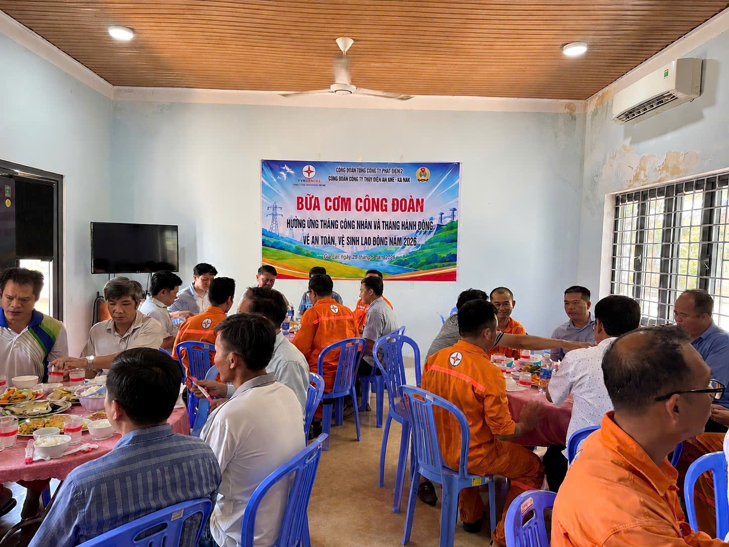 The image shows a group of people wearing orange shirts, gathered around a table, in a room with a large window and a noticeboard with text in Vietnamese, possibly a meeting or community event.AI-generated content may be incorrect.